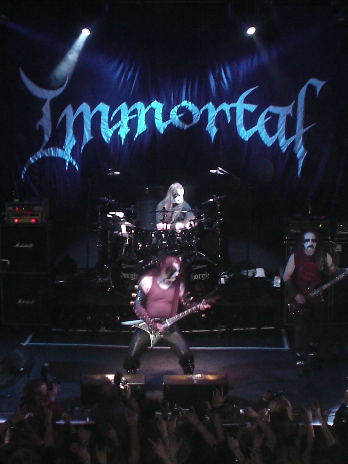 Músicos con la cara pintada de blanco y negro tocando bajo una luz azul, con el nombre Immortal en letras góticas en el telón