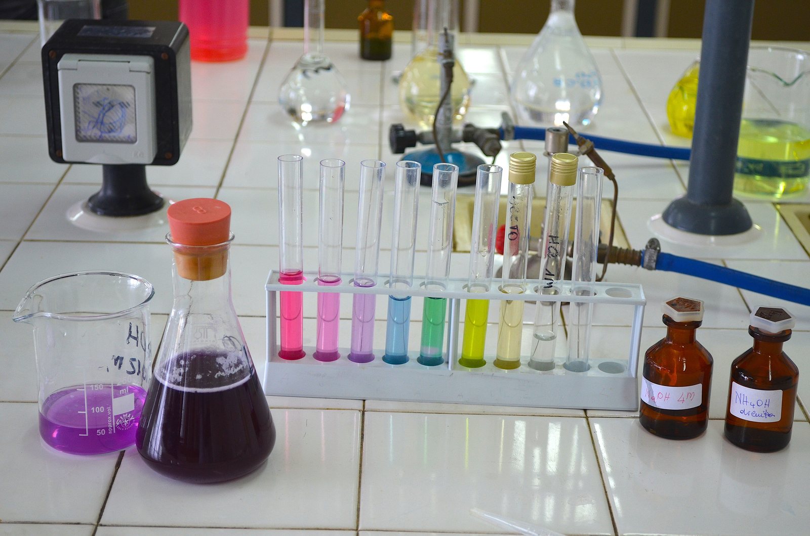 Tubos de ensayo con líquidos de todos los colores, del rosa al amarillo, junto a un matraz morado en un laboratorio